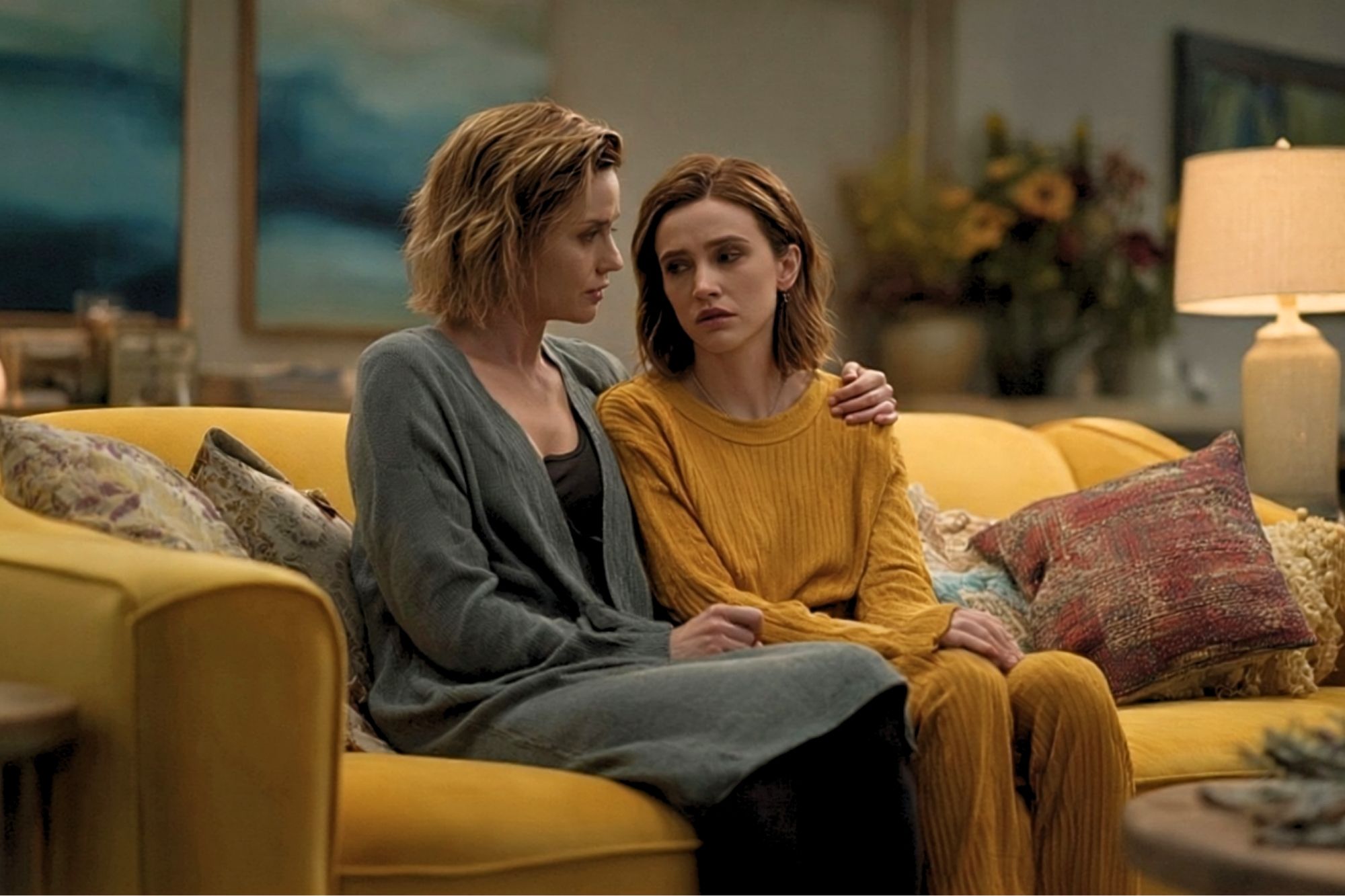 Two women sit close together on a yellow couch, one comforting the other with an arm around her shoulders. Both look serious and concerned. The room is warmly lit with art and flowers visible in the background.