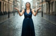 A woman in a sleeveless blue dress stands on a cobblestone street, smiling joyfully with arms raised, as rain pours down around her.