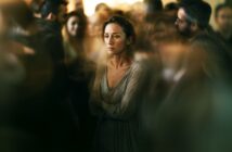 A woman stands alone in the middle of a blurred, crowded room, looking sad and distant. She has her arms crossed and gazes forward, highlighting a sense of isolation among the surrounding people.