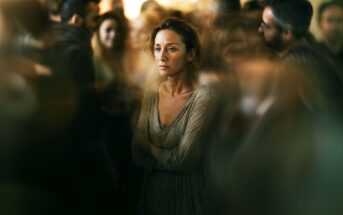 A woman stands alone in the middle of a blurred, crowded room, looking sad and distant. She has her arms crossed and gazes forward, highlighting a sense of isolation among the surrounding people.