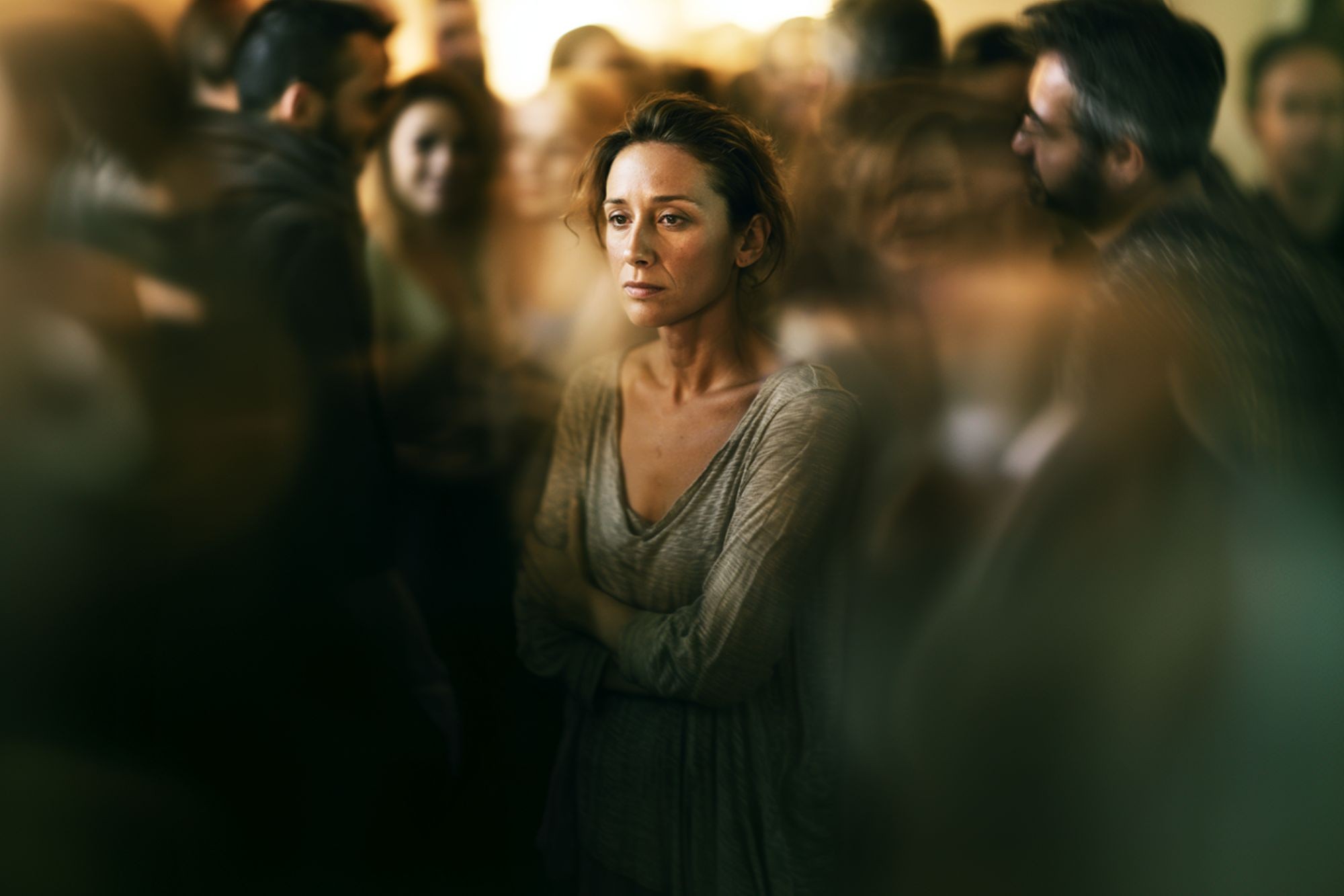 A woman stands alone in the middle of a blurred, crowded room, looking sad and distant. She has her arms crossed and gazes forward, highlighting a sense of isolation among the surrounding people.