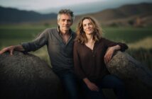 A man and woman sit close together on large rocks in an open field with mountains in the background, both smiling softly at the camera.