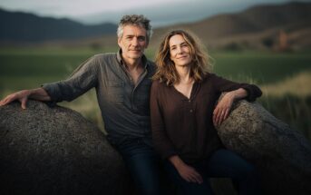 A man and woman sit close together on large rocks in an open field with mountains in the background, both smiling softly at the camera.