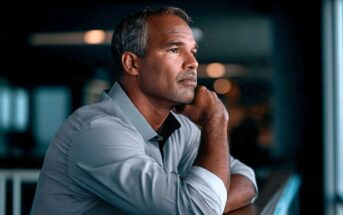 A man with short gray hair and a light blue shirt rests his chin on his hand, gazing thoughtfully into the distance in a softly lit indoor setting.