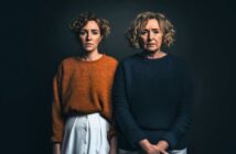 Two women with curly hair stand side by side against a dark background. The younger woman wears an orange sweater and white pants, while the older woman wears a dark blue sweater and light-colored pants. Both have serious expressions.