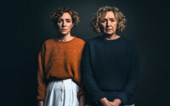 Two women with curly hair stand side by side against a dark background. The younger woman wears an orange sweater and white pants, while the older woman wears a dark blue sweater and light-colored pants. Both have serious expressions.
