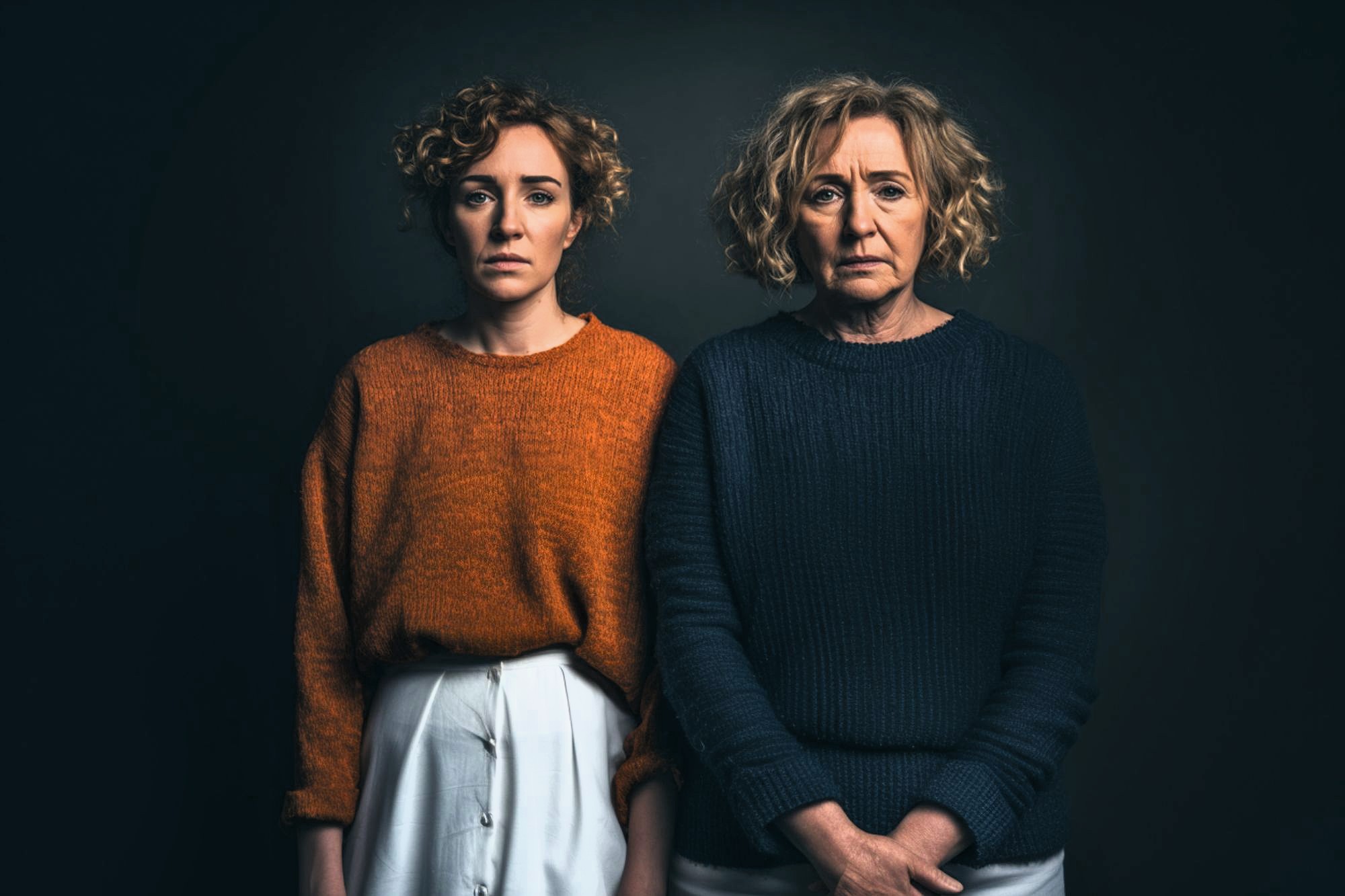Two women with curly hair stand side by side against a dark background. The younger woman wears an orange sweater and white pants, while the older woman wears a dark blue sweater and light-colored pants. Both have serious expressions.