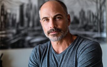 A middle-aged man with a beard and short hair wearing a gray shirt sits indoors, looking at the camera with a slight smile. A blurred black-and-white cityscape is in the background.