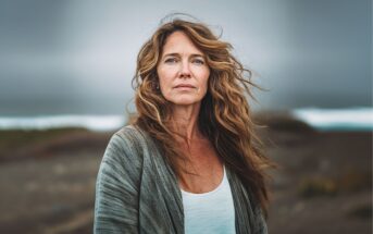 A woman with long wavy hair stands outdoors, wearing a gray cardigan over a white top. The background is blurred, showing a cloudy sky and what appears to be a beach or coastal landscape.
