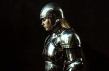 A person in shiny medieval armor and helmet stands in profile against a dark background, with long blond hair visible, looking downward in a contemplative pose.