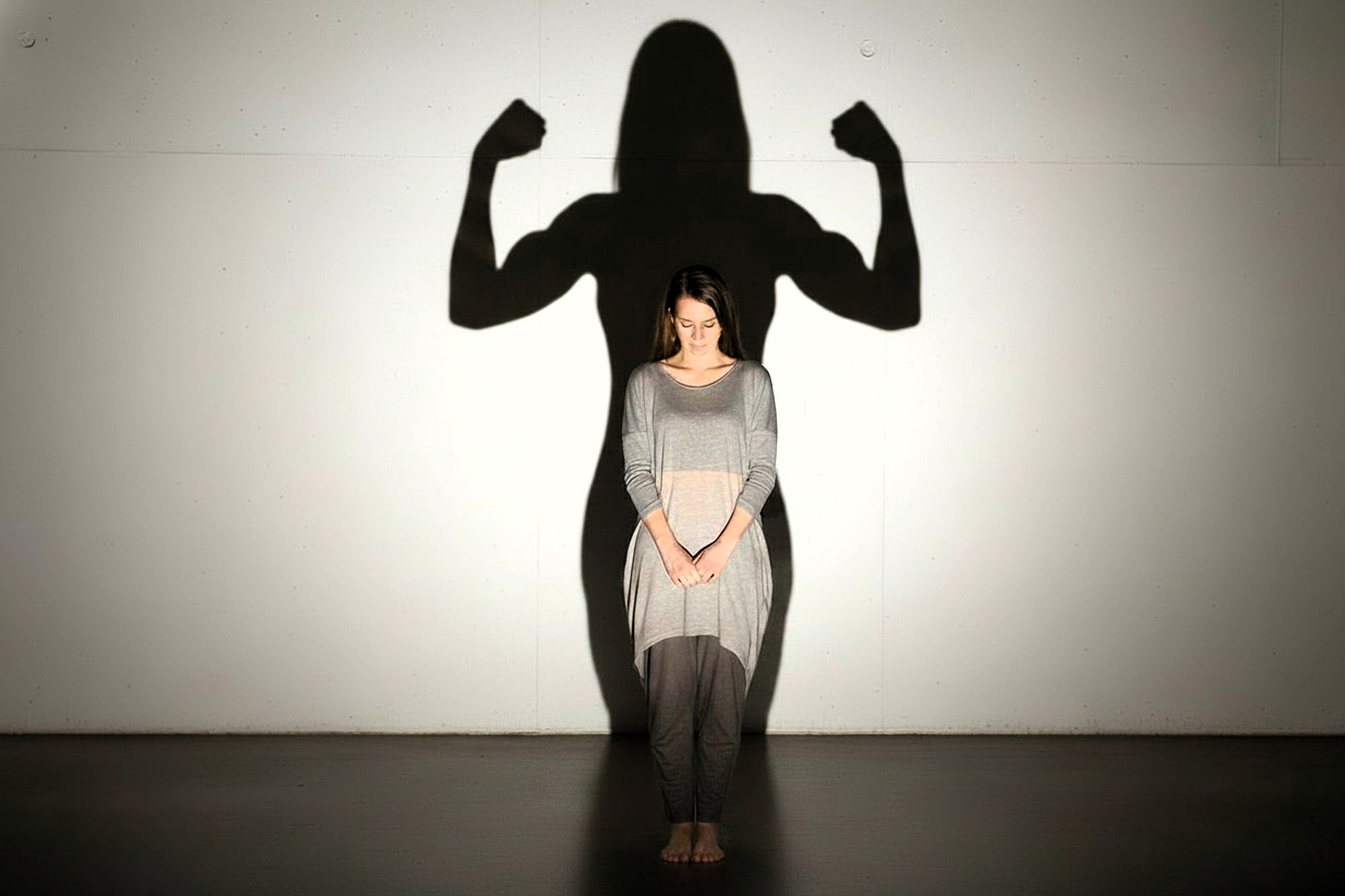 A woman stands barefoot with her head bowed, casting a shadow on the wall behind her that shows her in a strong, flexed-arm pose, symbolizing inner strength and confidence.