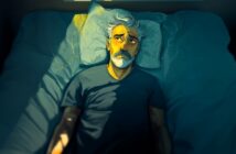 An older man with gray hair and a beard lies awake in bed at night, looking up thoughtfully. Soft yellow light from a window illuminates part of his face and pillow, contrasting with the dark, blue-toned room.