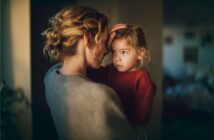 A woman with curly hair gently holds and comforts a young girl in a red sweater, placing her hand on the child’s head. The scene is softly lit and intimate, conveying warmth and care.