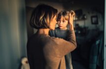 A woman gently holds a young girl's face, looking at her with concern. The child, wearing a gray sweater, looks back quietly. Soft, warm lighting creates an intimate and caring atmosphere in a cozy room.
