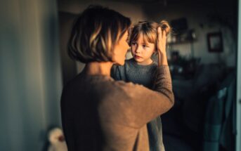 A woman gently holds a young girl's face, looking at her with concern. The child, wearing a gray sweater, looks back quietly. Soft, warm lighting creates an intimate and caring atmosphere in a cozy room.