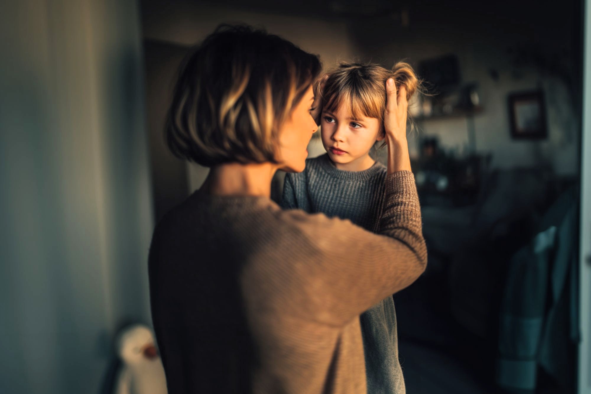 A woman gently holds a young girl's face, looking at her with concern. The child, wearing a gray sweater, looks back quietly. Soft, warm lighting creates an intimate and caring atmosphere in a cozy room.