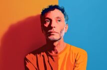 A man with curly hair and a beard, wearing an orange shirt, stands against a background split vertically into orange and blue halves, looking pensively to the side. The image has a stylized, digital illustration effect.