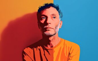 A man with curly hair and a beard, wearing an orange shirt, stands against a background split vertically into orange and blue halves, looking pensively to the side. The image has a stylized, digital illustration effect.