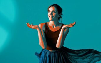 A woman in a sleeveless black top and flowing blue skirt stands against a blue background, smiling and gesturing upwards with both hands, as if expressing wonder or excitement.