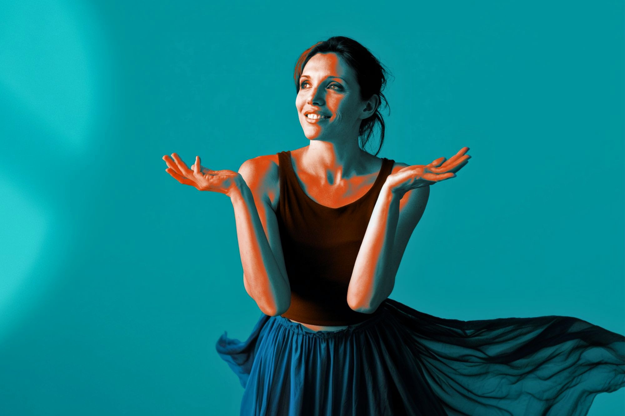 A woman in a sleeveless black top and flowing blue skirt stands against a blue background, smiling and gesturing upwards with both hands, as if expressing wonder or excitement.