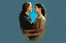 Two women stand closely, facing each other with serious expressions against a blue background. One has gray hair and wears a dark shirt; the other has dark hair and wears a light blouse. They gently hold each other’s arms.