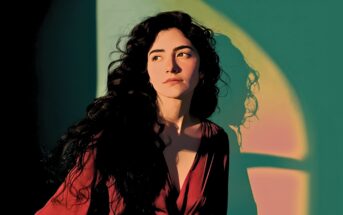 Woman with long curly hair in a deep red top stands in sunlight, casting a shadow on a teal wall with a rainbow arc. She looks thoughtfully to the side, her face illuminated by warm, dramatic light.