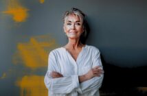 A confident older woman with short gray hair and glasses stands against a dark wall with orange paint streaks, wearing a loose white shirt and smiling gently with her arms crossed.