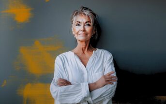 A confident older woman with short gray hair and glasses stands against a dark wall with orange paint streaks, wearing a loose white shirt and smiling gently with her arms crossed.