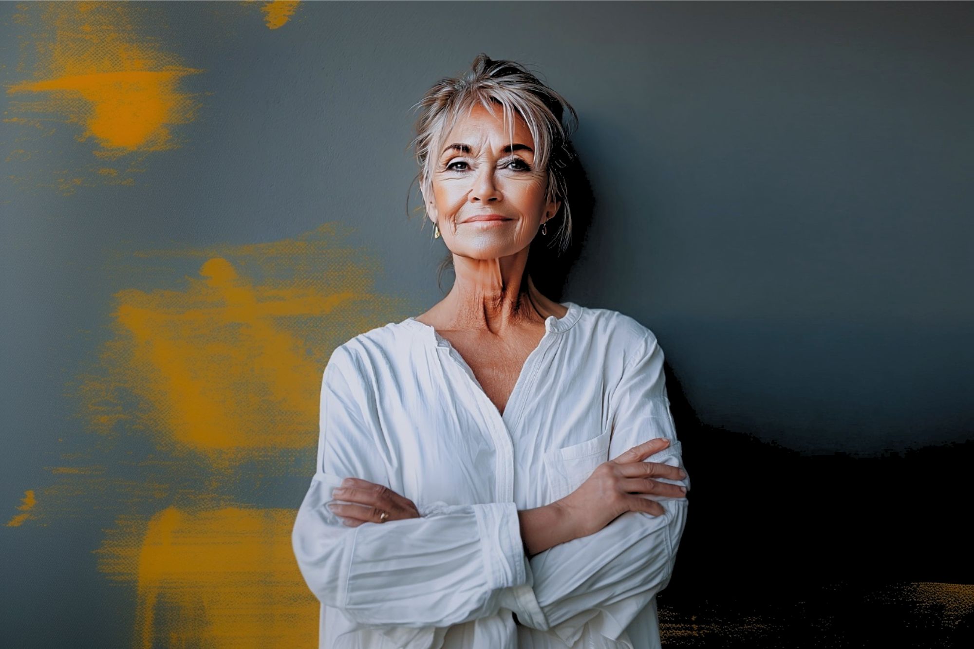 A confident older woman with short gray hair and glasses stands against a dark wall with orange paint streaks, wearing a loose white shirt and smiling gently with her arms crossed.