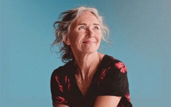 A smiling older woman with gray hair and a black shirt with red flowers looks upward against a plain blue background.