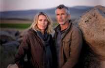 A middle-aged man and woman stand close together outdoors by large rocks, with open fields and hills in the background, both wearing jackets and looking calmly at the camera.