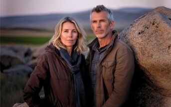 A middle-aged man and woman stand close together outdoors by large rocks, with open fields and hills in the background, both wearing jackets and looking calmly at the camera.