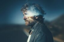 A man with tousled hair and a beard stands in profile, overlaid with a double exposure of a sunlit road leading toward distant mountains, blending his silhouette with the landscape.