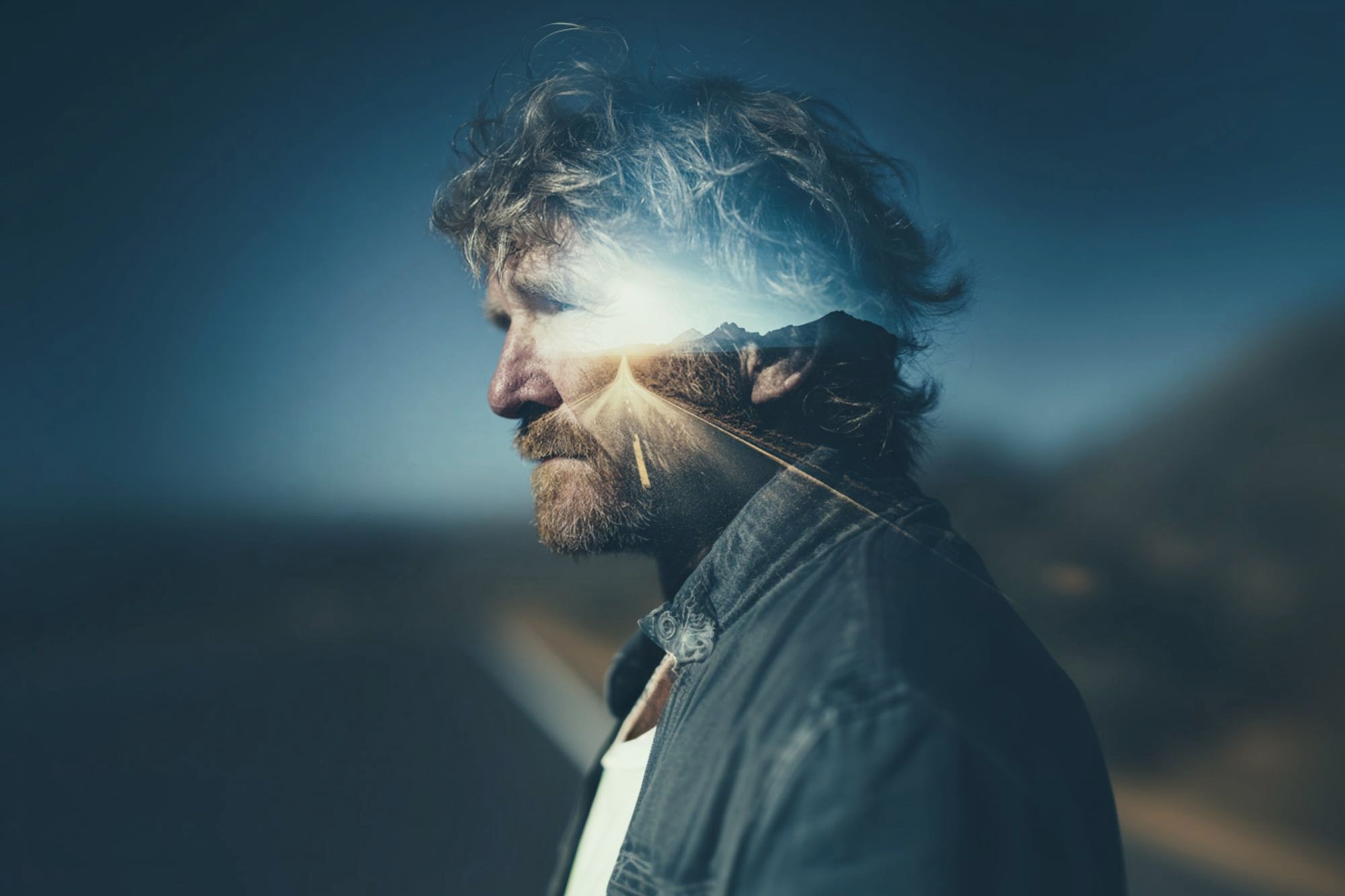 A man with tousled hair and a beard stands in profile, overlaid with a double exposure of a sunlit road leading toward distant mountains, blending his silhouette with the landscape.