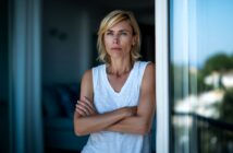 A woman with short blonde hair stands indoors near a window with her arms crossed, looking thoughtful and serious. She is wearing a white sleeveless top and natural light is coming in from outside.