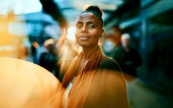 A woman stands outdoors in soft, golden sunlight, looking confidently at the camera. The background is blurred, with indistinct people and buildings, creating a warm, radiant atmosphere.