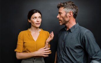 A woman in a yellow blouse gestures while talking to a man in a blue shirt against a dark background. They appear engaged in a serious conversation.