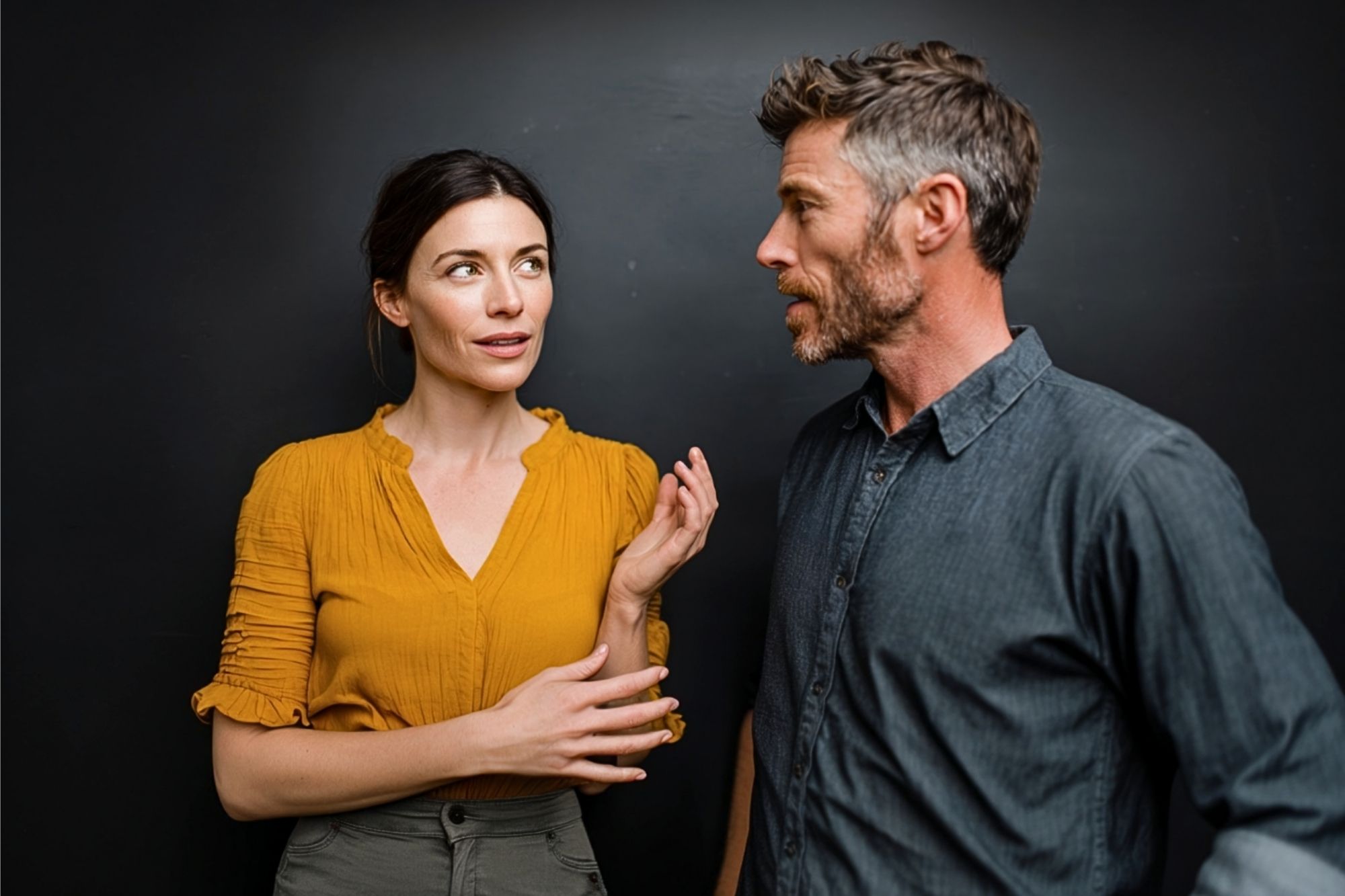A woman in a yellow blouse gestures while talking to a man in a blue shirt against a dark background. They appear engaged in a serious conversation.