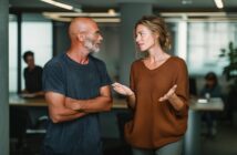 A man and a woman have a conversation in a modern office. The man is bald with glasses and a beard, wearing a dark t-shirt. The woman has light hair in a loose bun and wears a brown sweater, gesturing as she speaks.