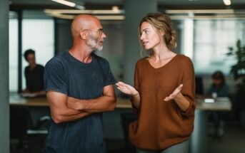 A man and a woman have a conversation in a modern office. The man is bald with glasses and a beard, wearing a dark t-shirt. The woman has light hair in a loose bun and wears a brown sweater, gesturing as she speaks.