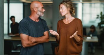 A man and a woman have a conversation in a modern office. The man is bald with glasses and a beard, wearing a dark t-shirt. The woman has light hair in a loose bun and wears a brown sweater, gesturing as she speaks.
