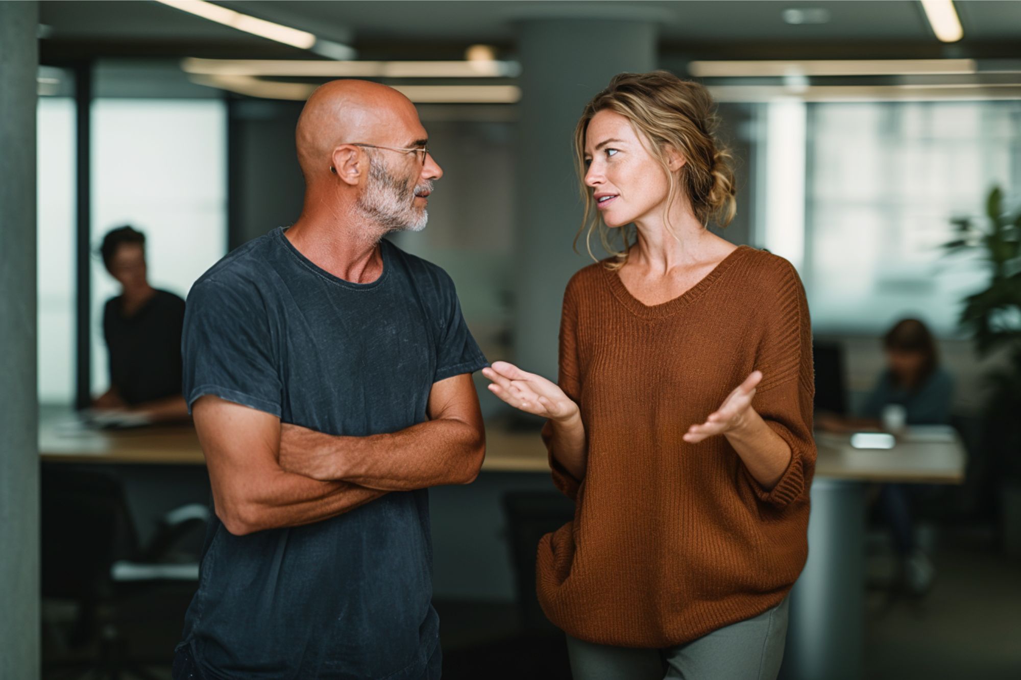 A man and a woman have a conversation in a modern office. The man is bald with glasses and a beard, wearing a dark t-shirt. The woman has light hair in a loose bun and wears a brown sweater, gesturing as she speaks.