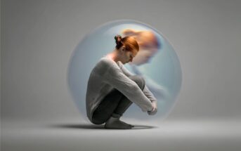 A woman with red hair sits curled up inside a transparent bubble, looking down. A blurred, ghostly duplicate of her appears behind, symbolizing isolation and emotional struggle. The background is plain gray.