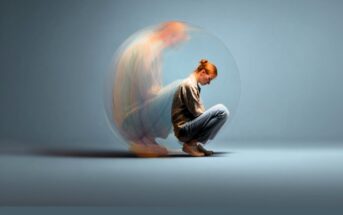 A young woman sits curled up on the floor with her head down, surrounded by a translucent sphere. A blurred, ghostly version of herself appears behind her, suggesting anxiety or emotional distress.