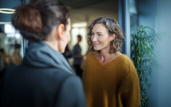 Two women stand facing each other indoors, engaged in conversation. One woman wears a mustard sweater, the other a dark coat. A green plant and blurred people are visible in the background.
