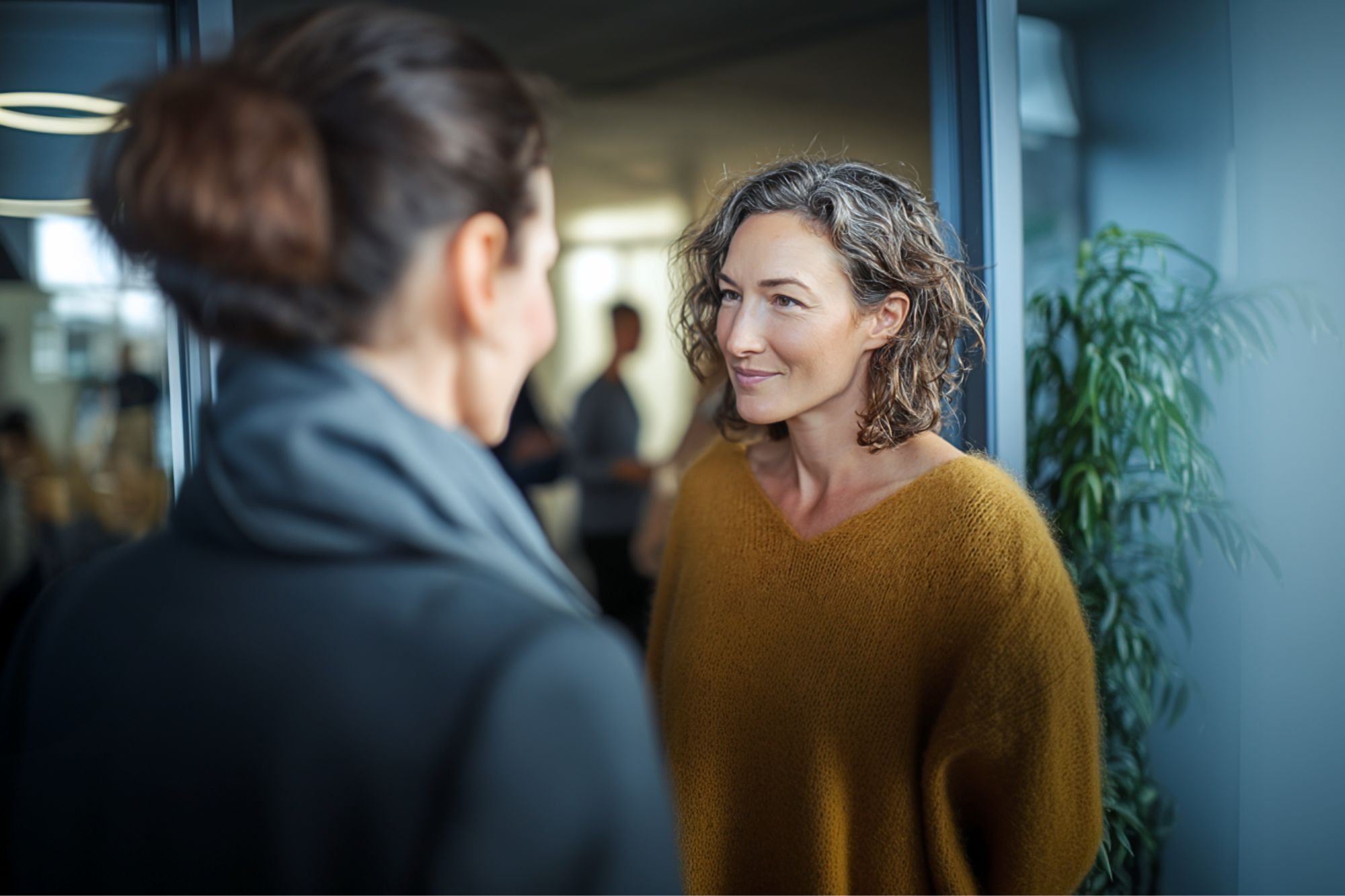 Two women stand facing each other indoors, engaged in conversation. One woman wears a mustard sweater, the other a dark coat. A green plant and blurred people are visible in the background.