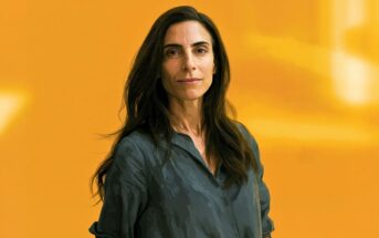 A woman with long brown hair wearing a loose gray shirt stands against a solid yellow background, looking confidently at the camera.