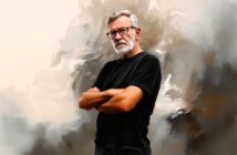 An older man with gray hair, a beard, and glasses stands with arms crossed, wearing a black t-shirt. The background is an abstract, painterly mix of neutral colors.