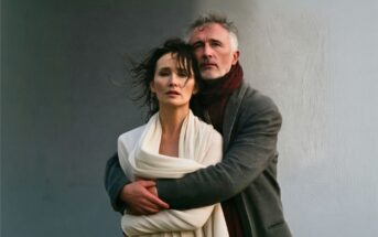 A middle-aged man in a gray coat hugs a woman draped in a white shawl. Both have serious expressions and stand against a plain, muted background.