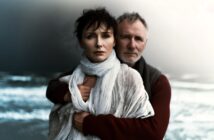 A woman wrapped in a white scarf stands in front of a man who embraces her from behind. Both look serious and pensive. The background is a blurred, moody seascape with waves and cloudy skies.